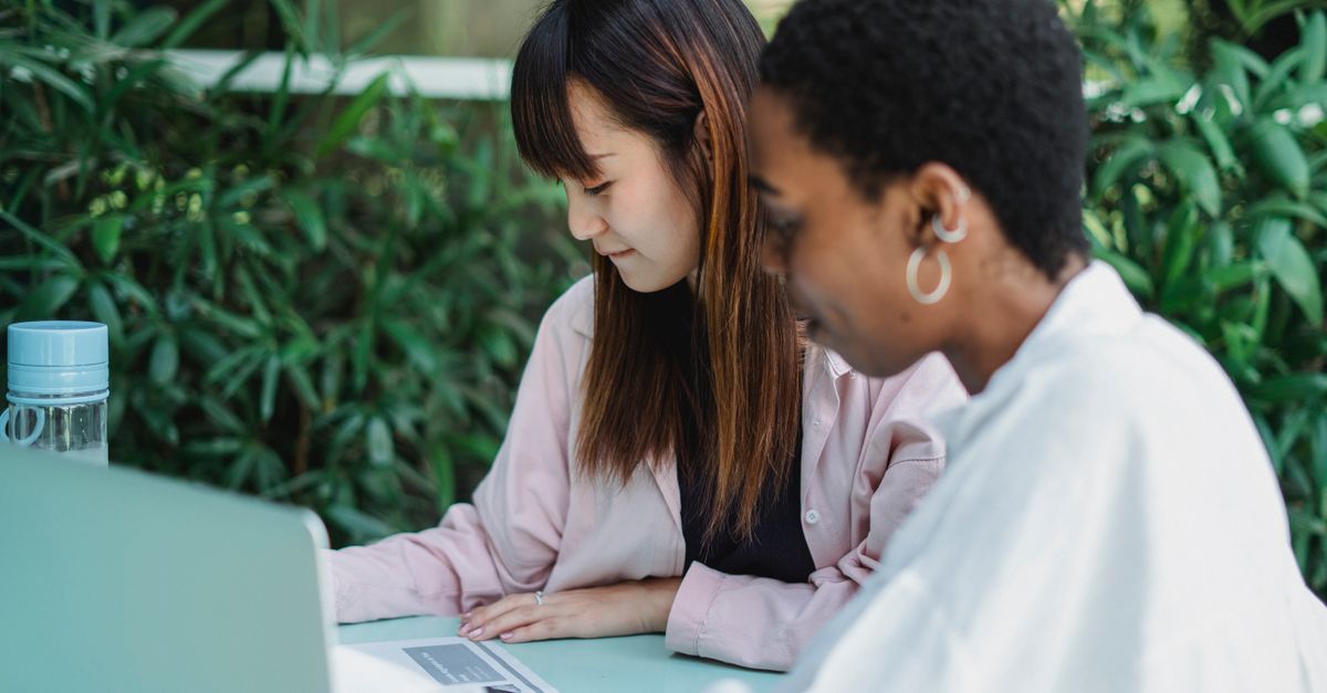 Two friends studying together outdoors with a laptop and notes.